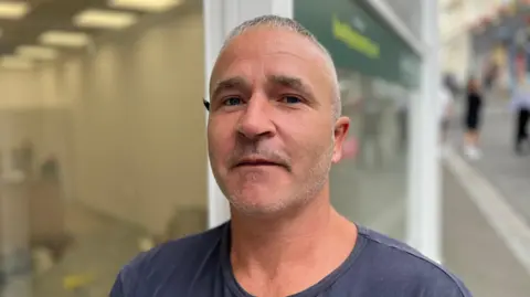 A man with close cropped grey hair and a pencil behind his right ear. He's wearing a blue t-shirt.