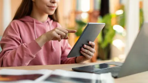 Pro-Stock Studio/Getty A girl in a pink hoodie sits at a desk in front of an open laptop. She holds an iPad in her left hand and points at the screen with her right index finger. A text book can be seen propped open next to the laptop.