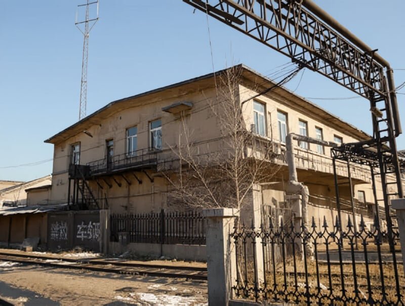 A two-story, beige building with peeling paint stands behind black iron fencing near railroad tracks, under a clear sky. Metal pipes and structures are visible around the building, and leafless trees grow nearby.