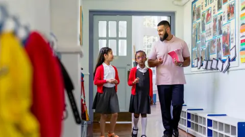 Getty Images A stock image of two female primary school pupils, both in red cardigans, walking along a school corridor with their male teacher who is wearing a pink polo shirt and carrying a stack of pink notebooks.