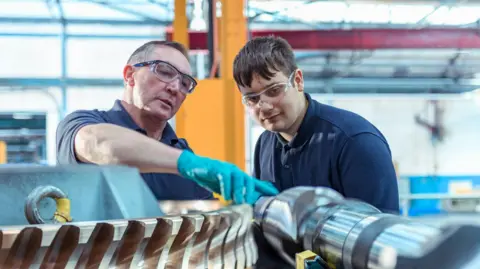 Getty Images An older man wearing blue plastic gloves is showing a large piece of metal equipment to a young man, both of whom are wearing short-sleeved navy polo shirts. They also have protective eye wear on and appear to be stood in a large factory warehouse, which is blurred in the background behind them.