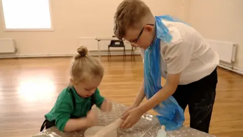 A teenage boy rolling out pizza dough with a toddler