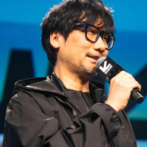 Getty Images Hideo Kojima. He has black hair in a short cut and wears large black glasses. He is smiling while he talks at a conference. He is wearing a trim black jacket and black t-shirt.