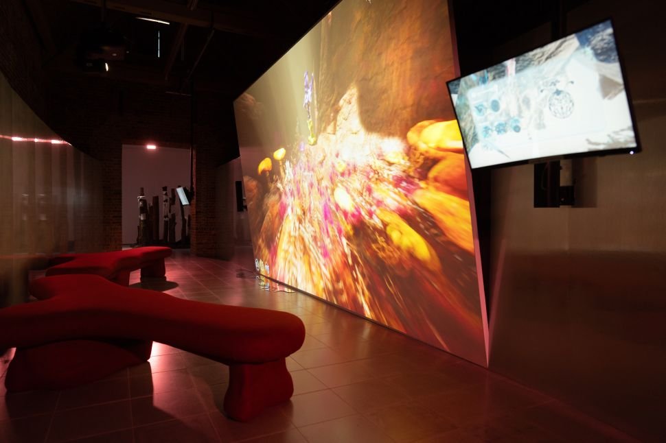 A darkened gallery room features large-scale immersive digital projections of glowing, abstract worlds with red sculptural seating in the foreground.