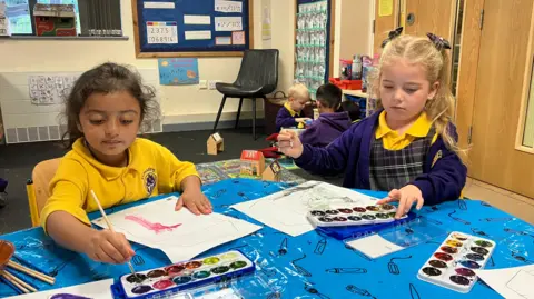 BBC/Kate McGough Two little girls, one asian with dark hair, one white with blonde hair sit at a table in reception class. The table is covered with a sheet and they are painting with brushes on a piece of paper. 