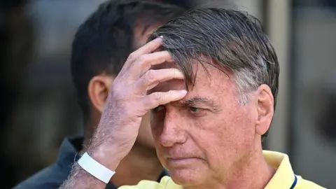 EVARISTO SA/AFP via Getty Images Former Brazilian President Jair Bolsonaro gestures after taking medical tests at DF Star hospital in Brasilia on August 16, 2025. He is wearing a pale yellow polo shirt and touches his forehead with his hand. 