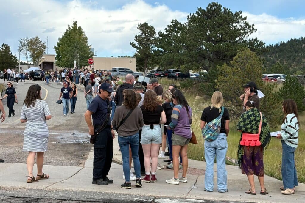 A long line of people stands and waits outside near trees and parked cars on a cloudy day. Some people talk to uniformed officers, while others look ahead or use their phones.