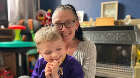 BBC/Kate McGough A woman in a grey top with long dark hair and glasses sits with her five year old son on her knee. They are both smiling.  